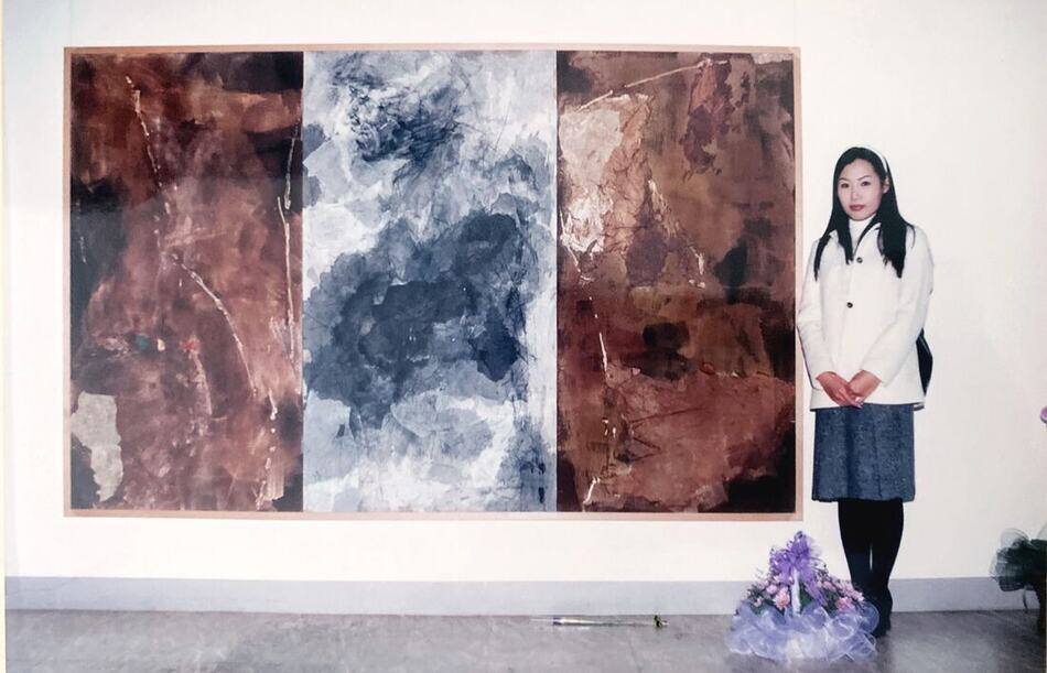 The artist standing beside a large three-panel abstract painting in earth reds and slate blue at her graduation exhibition
